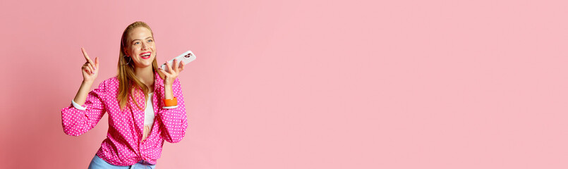 Banner. Happy attractive girl wearing pin up style clothes holding cell phone and recording voice message with cheerful facial expression over pink background.