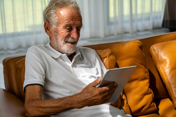 Caucasian old man smiling on the sofa Play tablet happily in the living room.