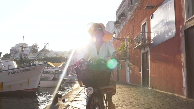 Young woman cycling with vintage bike along the harbour in Ponza island Italy. Fashion colorful dress skirt and hat.