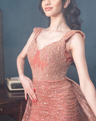 A woman in a pink dress stands in front of a vintage desk and looks into the camera.