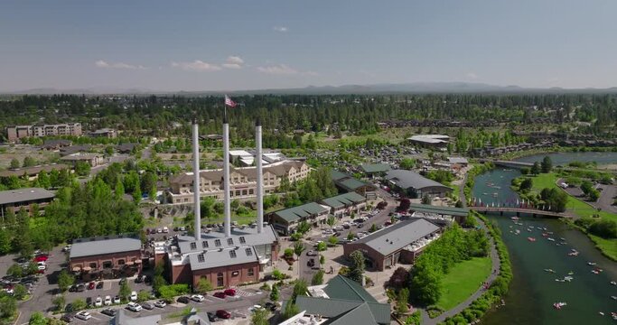 Aerial Forward Panning Shot Of American Flag On Industrial Pipe By Rowboats In River Amidst Green Landscape - Bend, Oregon