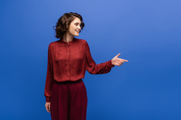 cheerful language teacher in maroon color outfit smiling while pointing with finger isolated on blue.