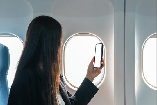A female traveler passenger sits at the window seat in economy class, using his smartphone, holding a mobile phone white screen mockup. close-up image