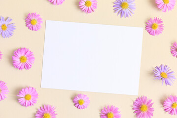 Greeting card template. Pink and purple flower aster on beige table background with blank paper sheet. Flat lay, top view, mockup, copy space for text. Aesthetic stylish floral pattern.