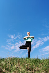 Woman stands on green grass against blue sky and doing yoga meditation exercise.