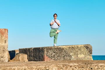 Happy tourist woman enjoying freedom while doing yoga on big stone at pier with blue sky in background.