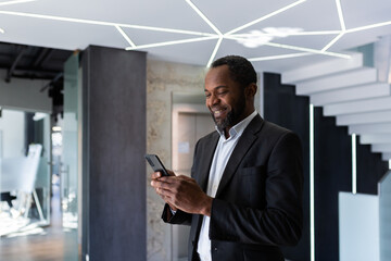 Successful african american businessman inside office standing by window, boss using phone, man typing message smiling and reading online news, typing message in social network app.