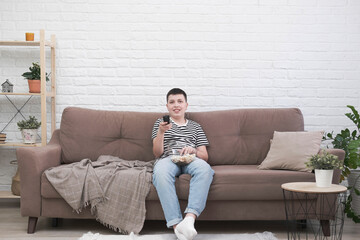 Obraz premium A teen boy is sitting on the sofa, eating pop corn, holding control panel and watching tv in light living room.