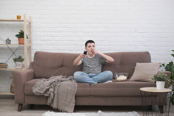 A teen boy is sitting on the sofa, eating pop corn, holding control panel and watching tv in light living room.