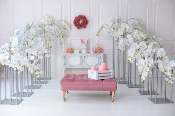 Elegance white living room interior with pink sofa pouf and decorative blossom flowers orchids. Wedding decor. Easter festive decoration with flowers wreath on wall, ceramic rabbits and box with eggs.