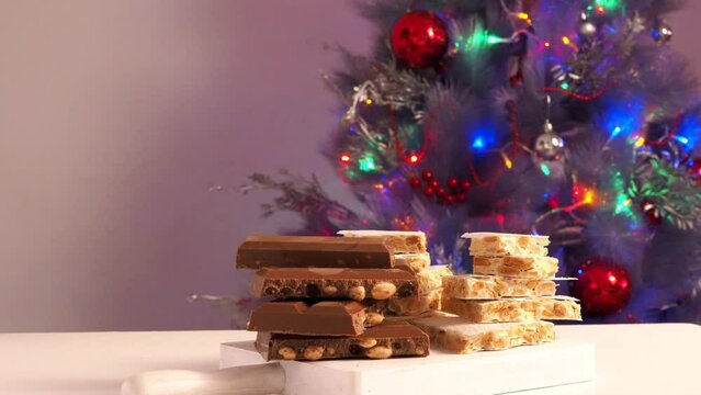Traditional Spanish Christmas sweets: turron with almonds and chocolate lying on a white board against the backdrop of a Christmas tree with a twinkling garland. 
