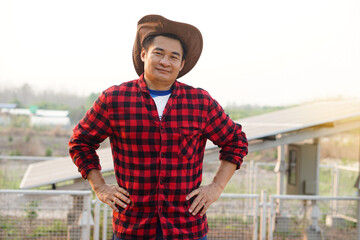 Handsome Asian man wears hat, red plaid shirt, stand beside solar panel at agriculture land, put hands on waists. feels confident. Concept, farmer installed photovoltaic for use in agriculture.