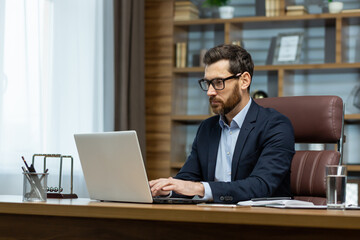 Successful mature businessman working inside office, man in business suit working at workplace with documents and laptop, boss behind paperwork attentive and focused.