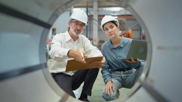 Two engineer manager leader and woman assistant holding laptop wearing helmet sitting at manufacturing factory discussing and explaining detail talking and checking production in workplace area