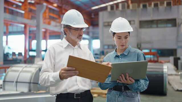 Two engineer manager leader and woman assistant holding laptop wearing helmet talking and checking production standing in workplace area at manufacturing factory