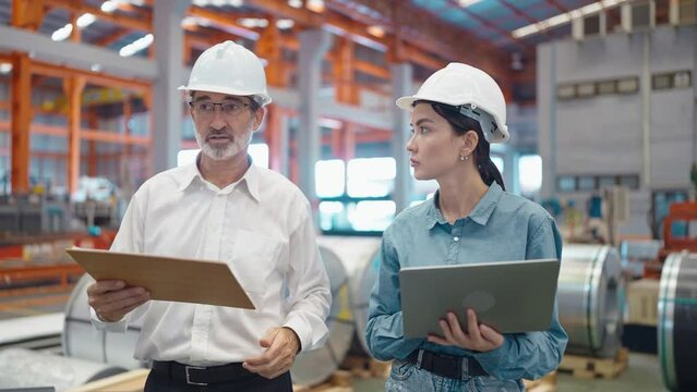 Two engineer manager leader and woman assistant holding laptop wearing helmet talking and checking production walking in workplace area at manufacturing factory