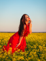 Beautiful young girl in a red dress posing in a field with canola. Happy woman on the nature. Spring season, warm day.