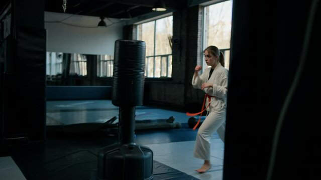 A girl in a kimono exercises with a punching bag in the gym while learning karate martial arts