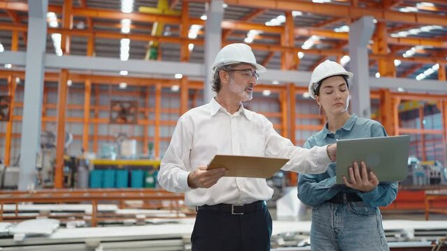 Two engineer manager leader and woman assistant holding laptop wearing helmet talking and checking production standing in workplace area at manufacturing factory