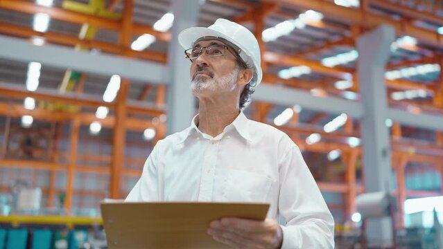 Engineer man manager leader wearing helmet holding clipboard standing and looking around at manufacturing factory recheck stock of production in workplace area