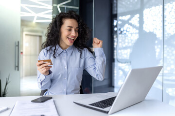 Financial success. A young Muslim businesswoman is sitting in the office at the table, looking at the laptop and holding a credit card in her hand. Rejoices, celebrates.