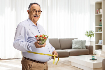 Mature man holding a salad and measuring waist in a living room