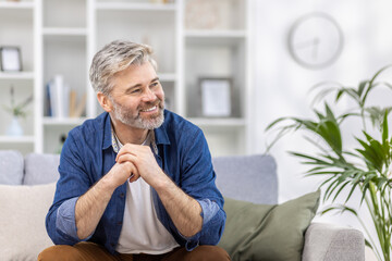 Portrait of a scary handsome gray-haired man in a blue shirt sitting at home on the sofa and smilingly looking to the side, crossing his arms in front of him.