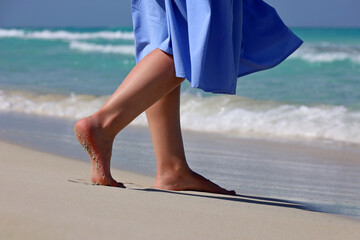 Barefoot woman in blue dress walking by the sand on sea waves background. Female legs on a coast, beach vacation - obrazy, fototapety, plakaty