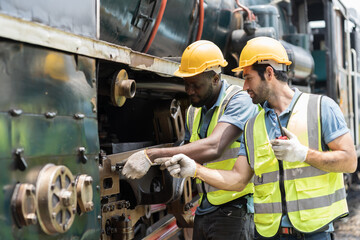 Two male engineer maintenance locomotive engine, wearing safety uniform, helmet and gloves in locomotive repair garage. Team male railway engineer use wrench repair train wheel in train garage
