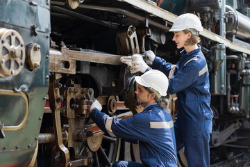 Male and female engineer maintenance locomotive engine, wearing safety uniform, helmet and gloves in locomotive repair garage. Male and female railway engineer use wrench repair train wheel in garage