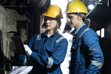 Male and female engineer maintenance locomotive engine, wearing safety uniform, helmet and gloves in locomotive garage. Male and female railway engineer use tablet while repair train wheel in garage