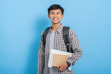 Young asian college student carrying books and backpack isolated over blue background
