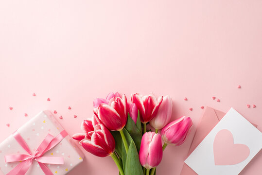 Mother's Day concept. Top view photo of bunch of pink tulips giftbox with bow envelope postal with heart and sprinkles on isolated pastel pink background with empty space