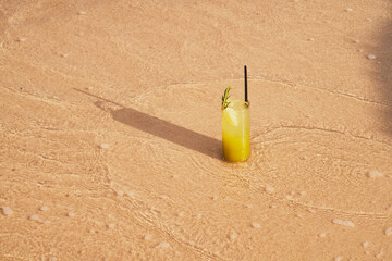 Image of a refreshing lemonade on the sand by the sea. Picture of cocktail among the waves by the sea at the beach. Visual evocative of a summer vacation trip in a tropical location.