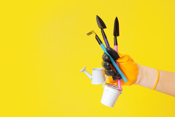 A hand in a glove holds gardening tools on a yellow background. Gardening concept