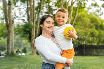 beautiful latina girl holding her son, while the boy holds a ball in his hands.