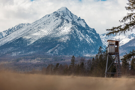 Snowy mountian Lomnicky stit from High tatras covered in snow and clouds while meadow under it is full of grass in a sunny day. Hunter hut or tower in the foreground.
