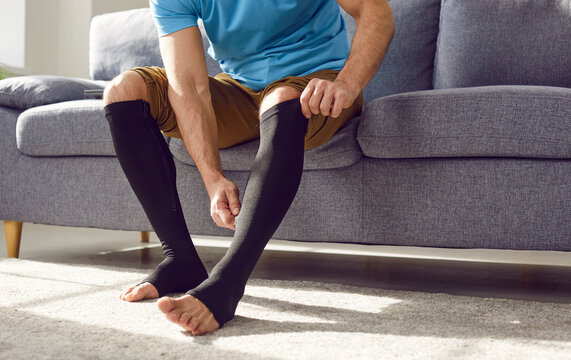 Cropped photo of a man putting on black medical compression stockings on his legs for the prevention of varicose veins and for venouse therapy sitting on the couch at home. Healthcare concept.