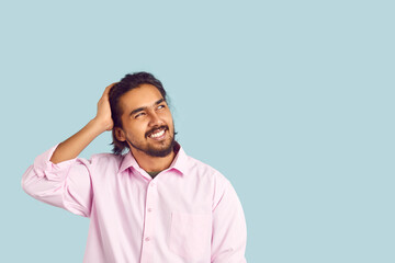 Man with funny facial expression is trying to remember what he forgot by looking at copy space. Portrait on pastel light blue background of confused Indian young man who doubts or made mistake. - obrazy, fototapety, plakaty