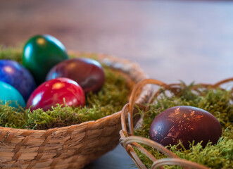 Various coloured decorated easter eggs in wicker basket nest with moss and colored Easter eggs on beige wooden bokeh background. For Happy Easter holiday card. Selective focus, copy space
