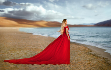 a beautiful blonde girl in a red long dress stands on a sandy seashore.