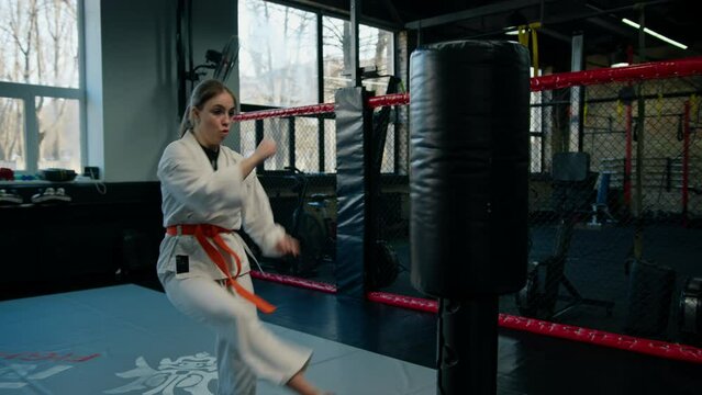 A girl in a kimono exercises with a punching bag in the gym while learning karate martial arts