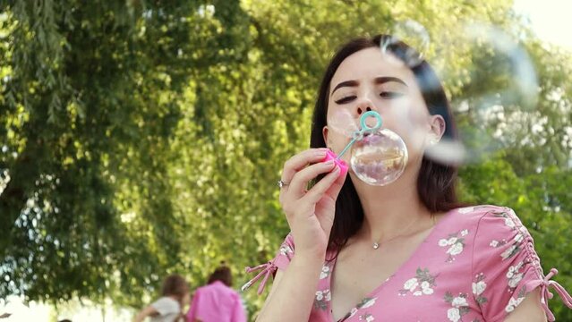 Woman sitting on white bedsheet, blowing soap bubbles, smiling. Lady have a rest in the city park, green grass lawn. Travel, vacation, picnic set
