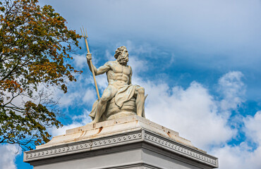Copenhagen, Denmark - September 13, 2010: Closeup, Neptunus or Poseidon God white stone statue on pillar at east side entrance to Churchill Parken. Blue cloudscape and green foliage