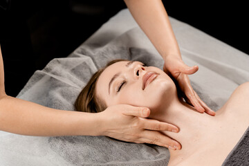 Girl with closed eyes is relaxing on face and neck massage in spa. Masseur is making facial massage for young woman in spa. Facebuilding and relaxing procedures.