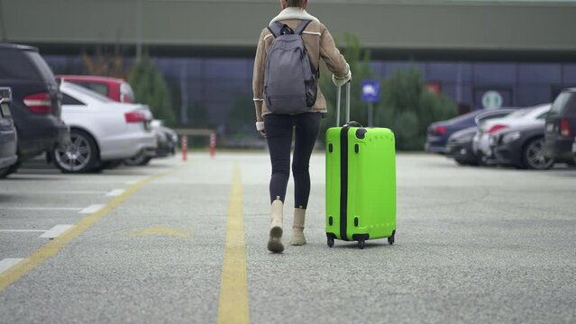 Traveler woman with backpack and horsetail hair walk pushing large suitcase on departure at airport, train station parking lot, slow motion cinematic shot