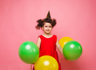 Portrait of a cheerful little girl isolated on a pink background, holding a bunch of colorful balloons, posing.