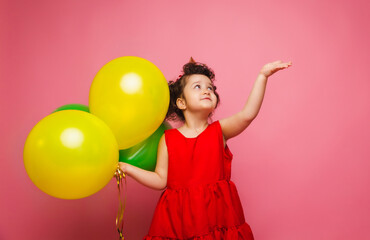Fototapeta na wymiar A joyful baby girl in a red dress celebrates her birthday and lets out colorful confetti on a pink background. a child holds balloons and catches confetti.