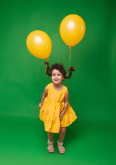 Fototapeta na wymiar Funny little girl on a green background. A girl is having fun with yellow balloons. balloons lift up the pigtails of the child.