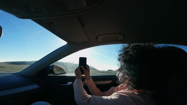 Commuter woman with curly hair taking pictures with phone from car traveling on mountain road at sunset